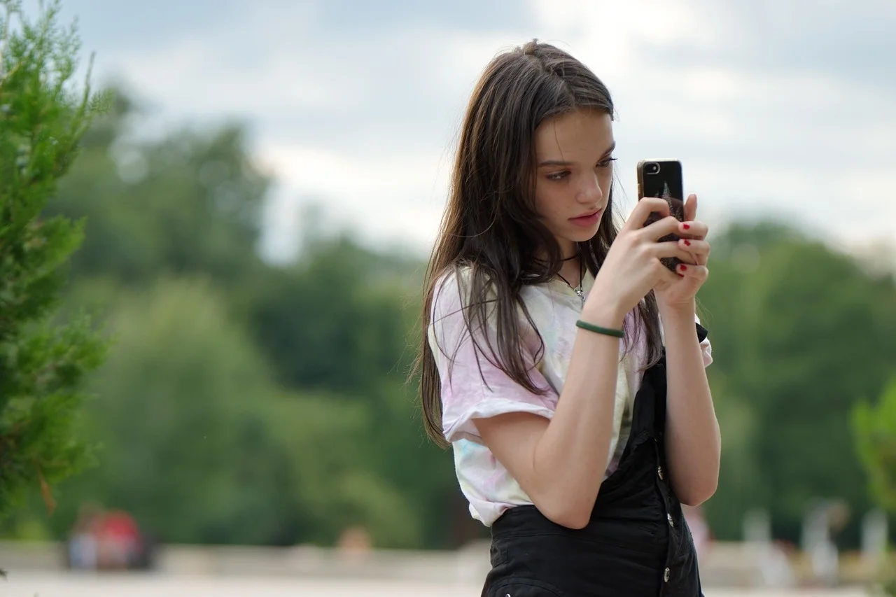 Eine junge Frau im Teenageralter hält ein Smartphone und fotografiert etwas im Freien in einem Park mit Bäumen.